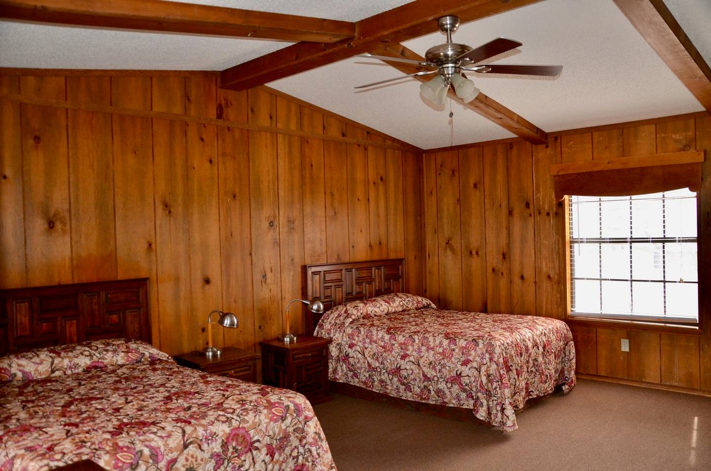 Inside a hillside cabin: two beds with floral spreads, knotty pine walls, a ceiling fan and a shuttered window