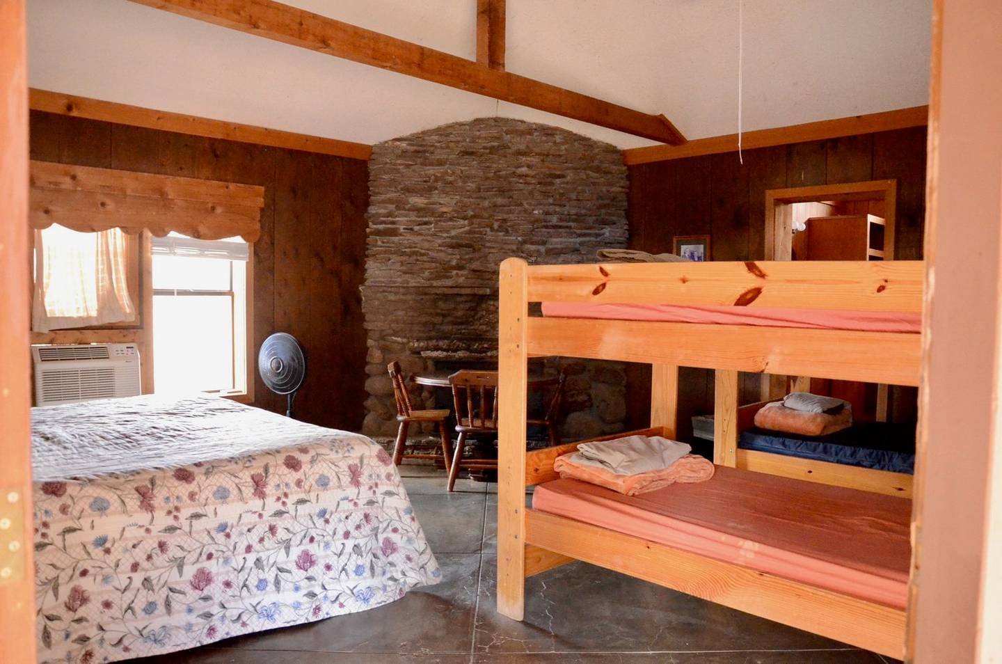 Inside a ranch cabin: a double bed and wooden bunk beds beside a stone fireplace, with a dining table by the window