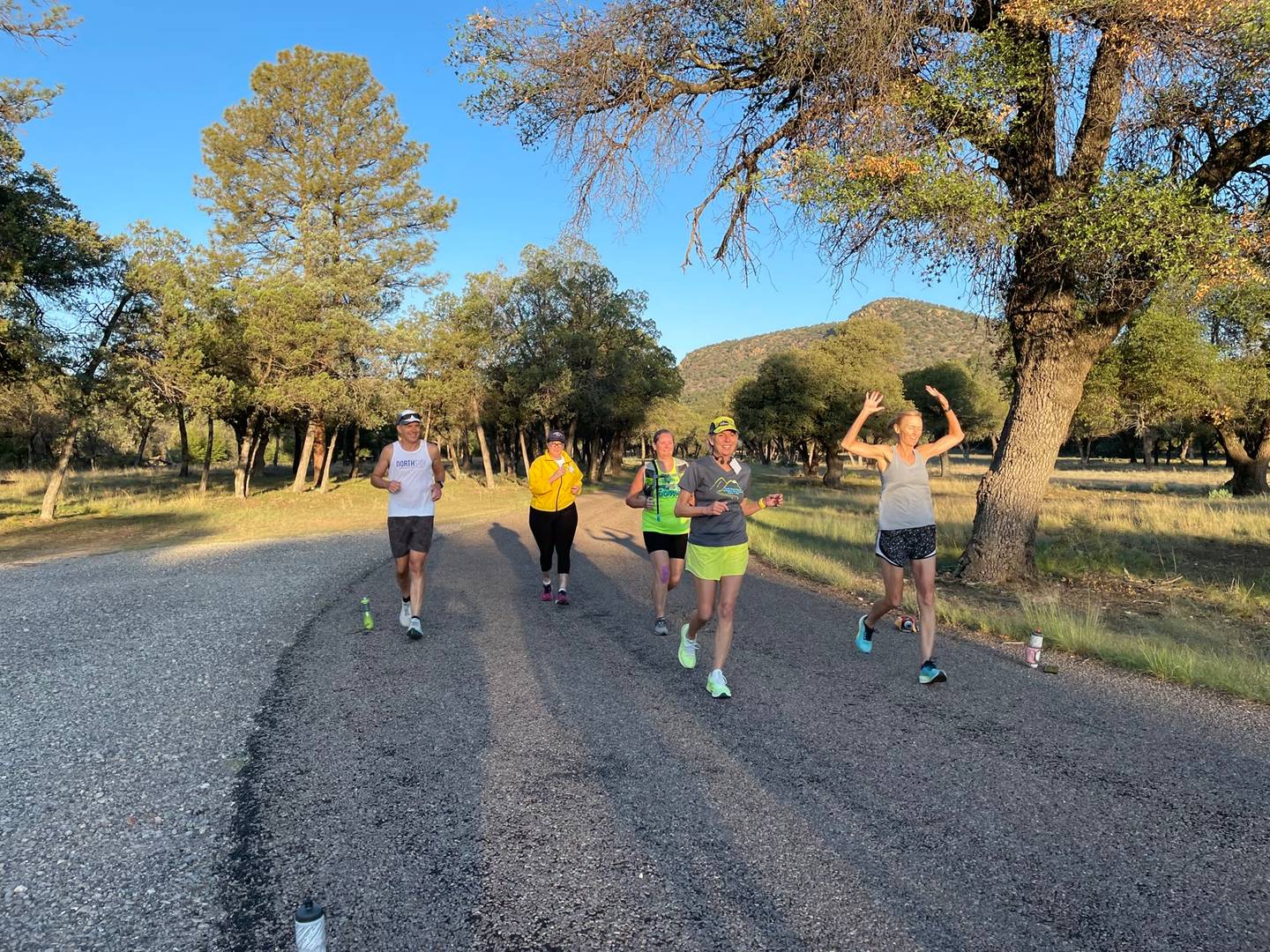 Five runners coming down a tree-lined ranch road in early morning light, one raising both arms