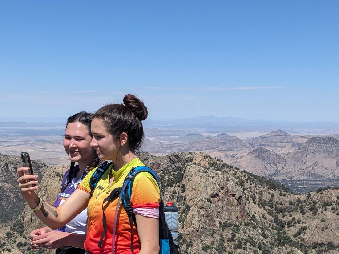 Two young hikers taking a phone photo at a summit with desert basin stretching to the horizon