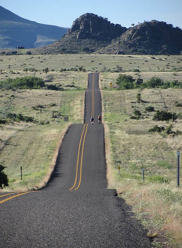 Two cyclists riding a rolling ribbon of empty road toward rocky desert peaks