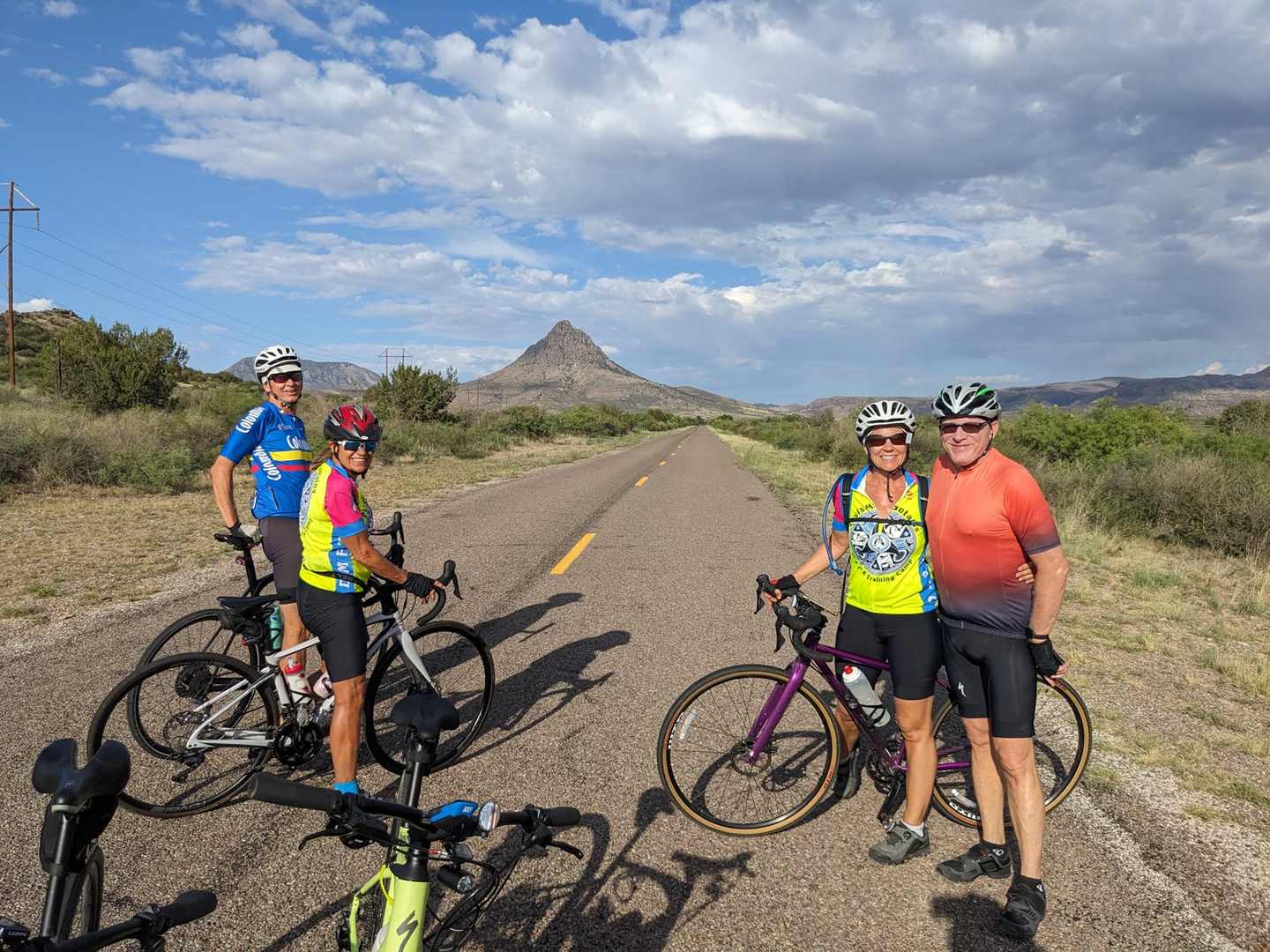 Four cyclists in helmets pausing on an empty two-lane road with a rocky peak behind them