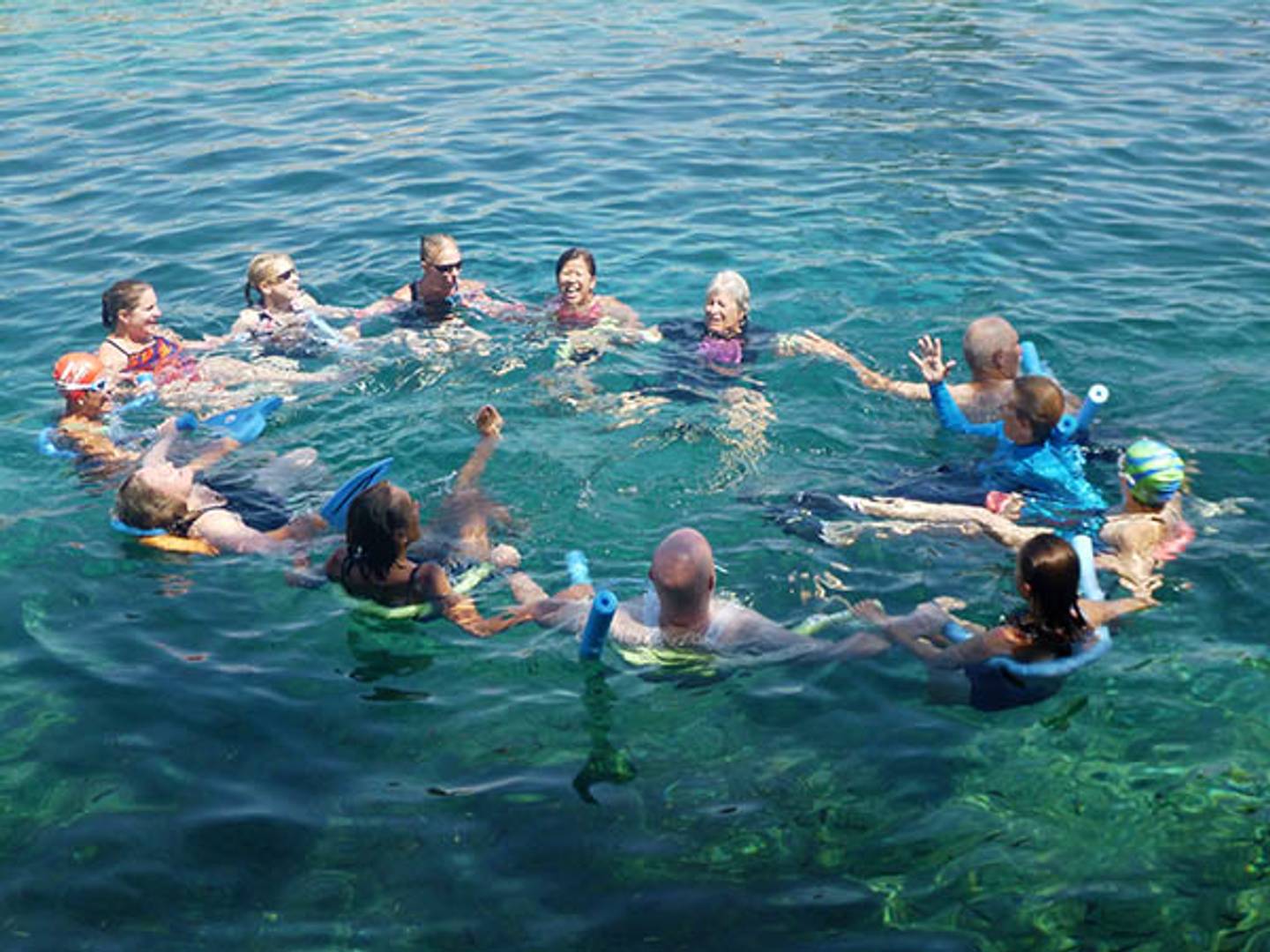 A dozen swimmers floating in a circle, holding pool noodles in clear spring water