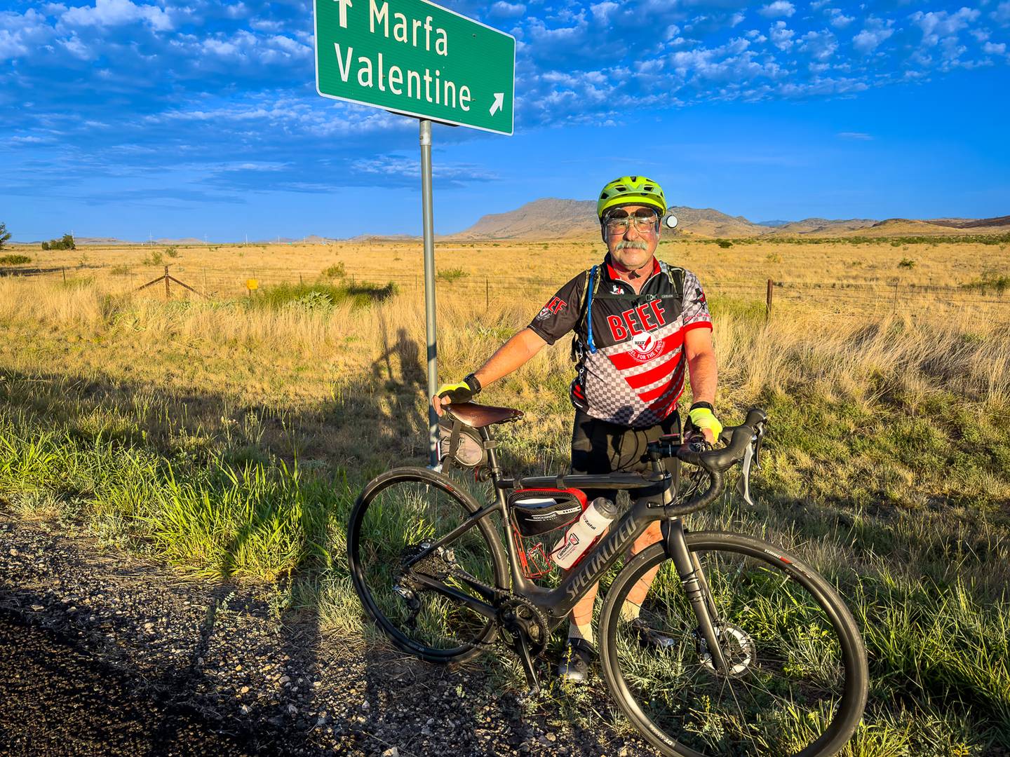 A smiling cyclist with a gray mustache standing with his bike under a Marfa and Valentine highway sign